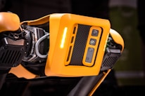 A detailed view of a robotic device featuring a bright orange casing with multiple cameras or sensors. The design includes perforated sections for ventilation and exposed mechanical parts, indicating advanced technology and engineering.