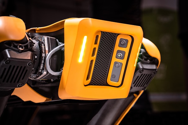 A detailed view of a robotic device featuring a bright orange casing with multiple cameras or sensors. The design includes perforated sections for ventilation and exposed mechanical parts, indicating advanced technology and engineering.