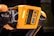 A detailed view of a robotic device featuring a bright orange casing with multiple cameras or sensors. The design includes perforated sections for ventilation and exposed mechanical parts, indicating advanced technology and engineering.