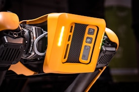 A detailed view of a robotic device featuring a bright orange casing with multiple cameras or sensors. The design includes perforated sections for ventilation and exposed mechanical parts, indicating advanced technology and engineering.