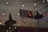 A string of warm lights decorates an outdoor space alongside hanging black witch hats. A vintage sign in the background advertises hotel accommodations, featuring phrases like 'clean' and 'reasonable'. A multi-story building with a partially open window is also visible.