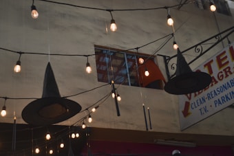 A string of warm lights decorates an outdoor space alongside hanging black witch hats. A vintage sign in the background advertises hotel accommodations, featuring phrases like 'clean' and 'reasonable'. A multi-story building with a partially open window is also visible.