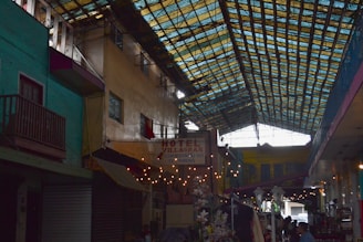 A covered marketplace or alleyway with vibrant, multicolored walls and a roof made of transparent panels allowing light to filter through. Several small businesses or kiosks line the sides, featuring goods and signage, including a sign for Hotel Villagran. Flowers and string lights add to the atmosphere, with people visible amidst the setting.