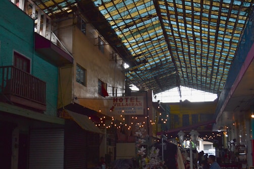 A covered marketplace or alleyway with vibrant, multicolored walls and a roof made of transparent panels allowing light to filter through. Several small businesses or kiosks line the sides, featuring goods and signage, including a sign for Hotel Villagran. Flowers and string lights add to the atmosphere, with people visible amidst the setting.