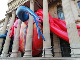 A large, colorful inflatable sculpture resembling a human heart is entwined around the classical columns of a historical building. The sculpture is primarily red and blue, with intricate vein patterns, creating a stark contrast against the traditional architectural elements of the stone building.
