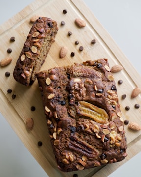 A loaf of banana bread topped with sliced bananas, almonds, walnuts, and chocolate chips is placed on a wooden cutting board. There are scattered whole almonds and chocolate chips around the bread, enhancing the rustic presentation.