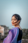 A woman is facing the camera with a gentle smile, wearing a traditional outfit with a vibrant purple and blue saree. Her hair is pinned back, and the background features a blurred view of houses and trees under a pale blue sky.