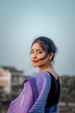 A woman is facing the camera with a gentle smile, wearing a traditional outfit with a vibrant purple and blue saree. Her hair is pinned back, and the background features a blurred view of houses and trees under a pale blue sky.