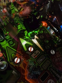 A pinball machine playfield with vibrant, colorful artwork. The design features a spooky theme with ghostly imagery, green hues, and illuminated elements. A small, glowing green figurine resembling a ghost or monster is present, adding to the eerie yet playful atmosphere.
