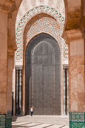Architectural close-up of an ornate, arched door embedded in a large stone facade with intricate geometric patterns. The door, adorned with floral and circular motifs, towers over a person standing in front, emphasizing its grand scale. The arch is heavily decorated with intricate mosaics featuring vibrant hues.