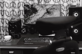 A home office desk setup featuring a computer monitor displaying a black and white image of waves. Surrounding the monitor are various audio equipment including speakers and a camera. A keyboard and mouse are placed on the desk surface, and a teacup sits on a coaster. A chair is partially visible in the foreground.