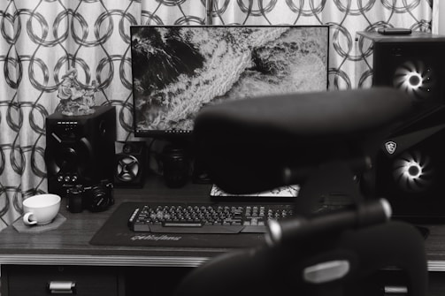 A home office desk setup featuring a computer monitor displaying a black and white image of waves. Surrounding the monitor are various audio equipment including speakers and a camera. A keyboard and mouse are placed on the desk surface, and a teacup sits on a coaster. A chair is partially visible in the foreground.
