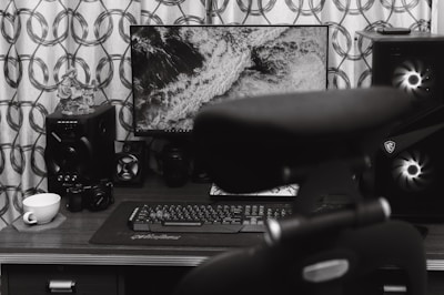 A home office desk setup featuring a computer monitor displaying a black and white image of waves. Surrounding the monitor are various audio equipment including speakers and a camera. A keyboard and mouse are placed on the desk surface, and a teacup sits on a coaster. A chair is partially visible in the foreground.