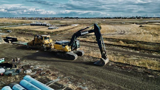 An expansive construction site features heavy machinery, including a large excavator and a bulldozer. The site is surrounded by open fields and some construction materials are scattered in the foreground. Piles of pipes are visible, and the sky is cloudy, suggesting an overcast day.