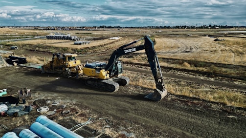 An expansive construction site features heavy machinery, including a large excavator and a bulldozer. The site is surrounded by open fields and some construction materials are scattered in the foreground. Piles of pipes are visible, and the sky is cloudy, suggesting an overcast day.