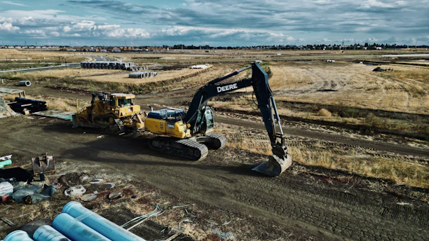 An expansive construction site features heavy machinery, including a large excavator and a bulldozer. The site is surrounded by open fields and some construction materials are scattered in the foreground. Piles of pipes are visible, and the sky is cloudy, suggesting an overcast day.