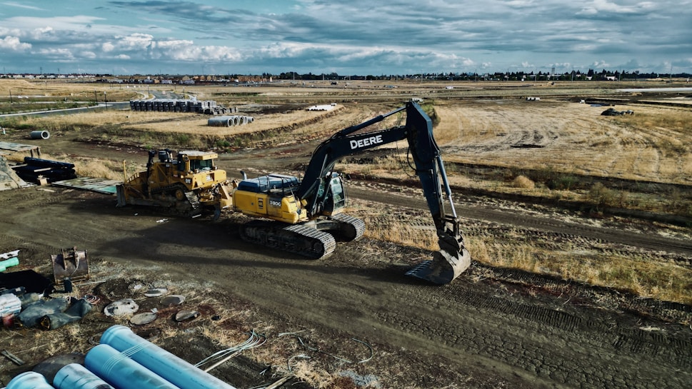 An expansive construction site features heavy machinery, including a large excavator and a bulldozer. The site is surrounded by open fields and some construction materials are scattered in the foreground. Piles of pipes are visible, and the sky is cloudy, suggesting an overcast day.