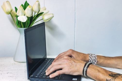 A person's hands are typing on a laptop placed on a white desk. A vase with white tulips is positioned next to the laptop. The person is wearing bracelets and has a tattoo on their arm. The background is a plain light-colored wall, and the overall setting appears to be minimalist and tidy.