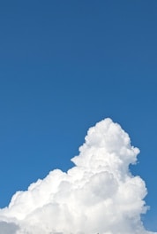 A large, fluffy cumulus cloud rises against a clear blue sky. The cloud is bright white, with billowing layers that give it a soft, pillowy appearance.