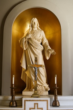 A statue depicting a religious figure in flowing robes with an extended arm, set against a golden niche. Two ornate candlesticks and a crucifix are positioned in front of the statue, creating a sense of reverence.