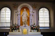 An ornate altar with a statue of a religious figure, surrounded by two stained glass windows. The altar is adorned with flowers and a decorative cross. The words 'Virgo Fidelis' are inscribed above the statue, and intricate gold and marble details embellish the setting.