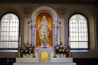 An ornate altar with a statue of a religious figure, surrounded by two stained glass windows. The altar is adorned with flowers and a decorative cross. The words 'Virgo Fidelis' are inscribed above the statue, and intricate gold and marble details embellish the setting.