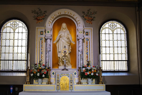 An ornate altar with a statue of a religious figure, surrounded by two stained glass windows. The altar is adorned with flowers and a decorative cross. The words 'Virgo Fidelis' are inscribed above the statue, and intricate gold and marble details embellish the setting.