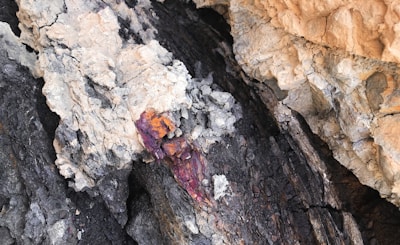 A rugged rock face with layers of dark and light minerals. There are veins of vibrant reddish-orange and purple hues cutting through the rock, indicating the presence of different mineral compositions.