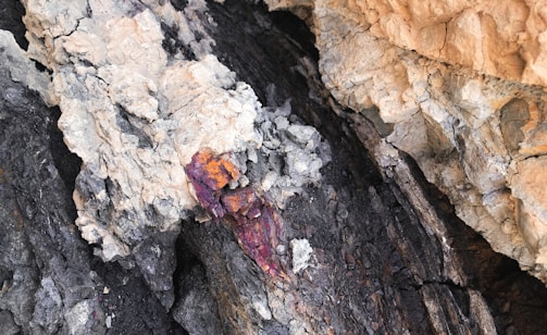 A rugged rock face with layers of dark and light minerals. There are veins of vibrant reddish-orange and purple hues cutting through the rock, indicating the presence of different mineral compositions.