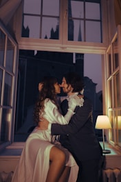 A couple is intimately sitting together by an open window, embracing each other with affectionate smiles. They are framed by the dim, warm glow of a lamp and the dark night sky outside. Both are dressed elegantly, with the woman in a silky gown and the man in a formal suit, suggesting a romantic or celebratory atmosphere.
