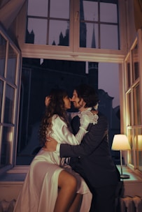 A couple is intimately sitting together by an open window, embracing each other with affectionate smiles. They are framed by the dim, warm glow of a lamp and the dark night sky outside. Both are dressed elegantly, with the woman in a silky gown and the man in a formal suit, suggesting a romantic or celebratory atmosphere.