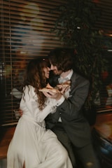 A couple sitting closely together indoors, sharing slices of pizza with a backdrop of blinds and a leafy plant. Both are dressed elegantly, with the woman in a long white dress and the man in a dark suit. They appear happy and intimate.