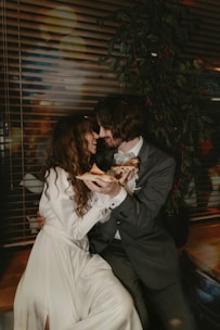 A couple sitting closely together indoors, sharing slices of pizza with a backdrop of blinds and a leafy plant. Both are dressed elegantly, with the woman in a long white dress and the man in a dark suit. They appear happy and intimate.