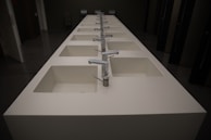 A row of modern, white rectangular sinks with metallic faucets extending along the length of a countertop. The setting appears to be a public restroom with dark walls and doors visible in the background.