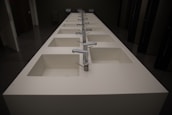 A row of modern, white rectangular sinks with metallic faucets extending along the length of a countertop. The setting appears to be a public restroom with dark walls and doors visible in the background.