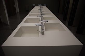 A row of modern, white rectangular sinks with metallic faucets extending along the length of a countertop. The setting appears to be a public restroom with dark walls and doors visible in the background.