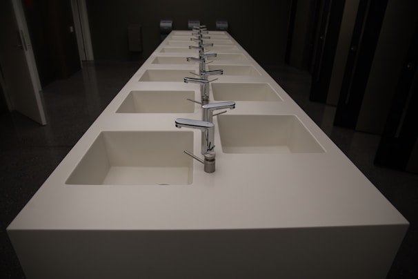 A row of modern, white rectangular sinks with metallic faucets extending along the length of a countertop. The setting appears to be a public restroom with dark walls and doors visible in the background.