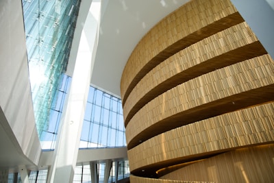 A modern architectural interior featuring a large, curved wooden structure with layered horizontal slats. The surrounding space is filled with natural light streaming through the tall glass windows. The high ceiling and angular walls contribute to an open and airy atmosphere.