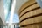A modern architectural interior featuring a large, curved wooden structure with layered horizontal slats. The surrounding space is filled with natural light streaming through the tall glass windows. The high ceiling and angular walls contribute to an open and airy atmosphere.