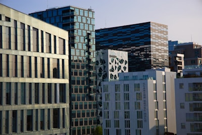 A cluster of modern office buildings with varying architectural designs, featuring a mix of glass, concrete, and geometric patterns. The buildings are closely packed, with one structure prominently displaying reflective glass panels, contrasting sharply with the darker, striped facade of another. The scene is set against a clear sky, suggesting a typical urban cityscape.