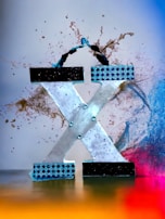 A large, stylized letter 'X' is decorated with a combination of black and blue patterned elements. The background features dynamic splashes of brown, gradually fading into blue and then pink, with a base of golden yellow tones, creating a vibrant and energetic atmosphere.