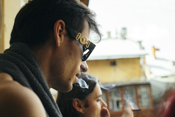 A profile view of a person smoking, wearing dark sunglasses and a towel draped around the neck. Smoke is visible as it rises from the cigarette in the person's hand. The background features an out-of-focus view of a building with a distinctively urban setting.