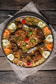 A large platter of biryani featuring grilled chicken pieces topped with herbs and garnished with cashew nuts. Surrounding the biryani are decorative slices of cucumber, carrot, red onion, and lemon, along with artistic rosettes made of tomato peels. The presentation is on a rustic wooden table covered with a cloth.