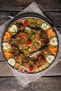A large platter of biryani featuring grilled chicken pieces topped with herbs and garnished with cashew nuts. Surrounding the biryani are decorative slices of cucumber, carrot, red onion, and lemon, along with artistic rosettes made of tomato peels. The presentation is on a rustic wooden table covered with a cloth.
