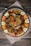 A large platter of biryani featuring grilled chicken pieces topped with herbs and garnished with cashew nuts. Surrounding the biryani are decorative slices of cucumber, carrot, red onion, and lemon, along with artistic rosettes made of tomato peels. The presentation is on a rustic wooden table covered with a cloth.