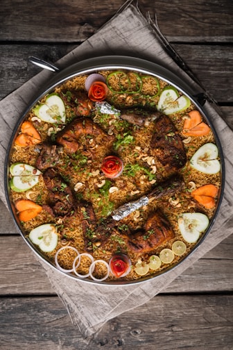 A large platter of biryani featuring grilled chicken pieces topped with herbs and garnished with cashew nuts. Surrounding the biryani are decorative slices of cucumber, carrot, red onion, and lemon, along with artistic rosettes made of tomato peels. The presentation is on a rustic wooden table covered with a cloth.