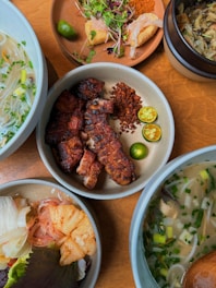A variety of dishes arranged on a wooden table. The central plate features grilled meat garnished with sliced calamansi and chili flakes. Surrounding dishes include noodle soup with herbs, a bowl of pickled vegetables, and a plate with fresh greens and lime. A small pot contains what appears to be vegetable or mushroom stew.