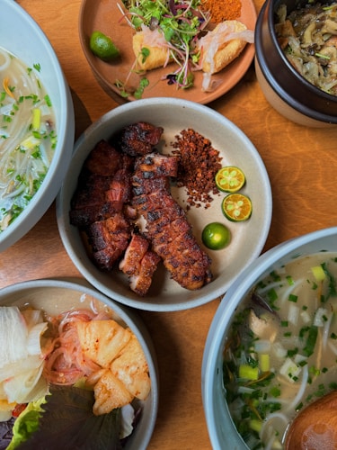 A variety of dishes arranged on a wooden table. The central plate features grilled meat garnished with sliced calamansi and chili flakes. Surrounding dishes include noodle soup with herbs, a bowl of pickled vegetables, and a plate with fresh greens and lime. A small pot contains what appears to be vegetable or mushroom stew.