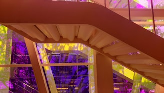 A modern staircase with a sleek design is positioned against a backdrop of colorful tinted glass panels. The vibrant hues of purple and yellow create an abstract effect with shadows and reflections.