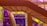 A modern staircase with a sleek design is positioned against a backdrop of colorful tinted glass panels. The vibrant hues of purple and yellow create an abstract effect with shadows and reflections.